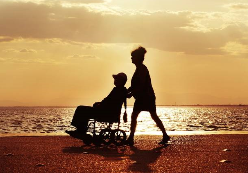 Caregiver spinge persona con disabilità in carrozzina. Sullo sfondo mare e cielo giallo