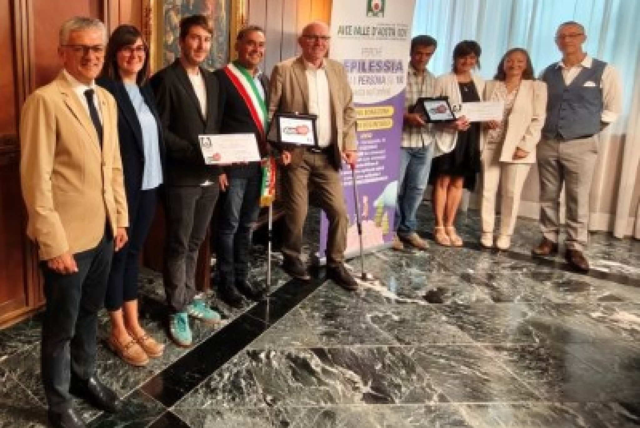 La consegna del premio dell'AICE "6con noi". Da sinistra l’assessore regionale ai Trasporti Luigi Bertschy; l’assessora al Turismo di Doues Corinne Cuaz; il vicesindaco di Doues Daniel Diemoz; il Sindaco di Doues Giorgio Abram; il presidente della Regione Renzo Testolin; i genitori della minore di Doues; la coordinatrice dell’Istituzione Scolastica Grand Combin Simona d’Agostino; il presidente dell'AICE Valle d’Aosta Manuele Amateis