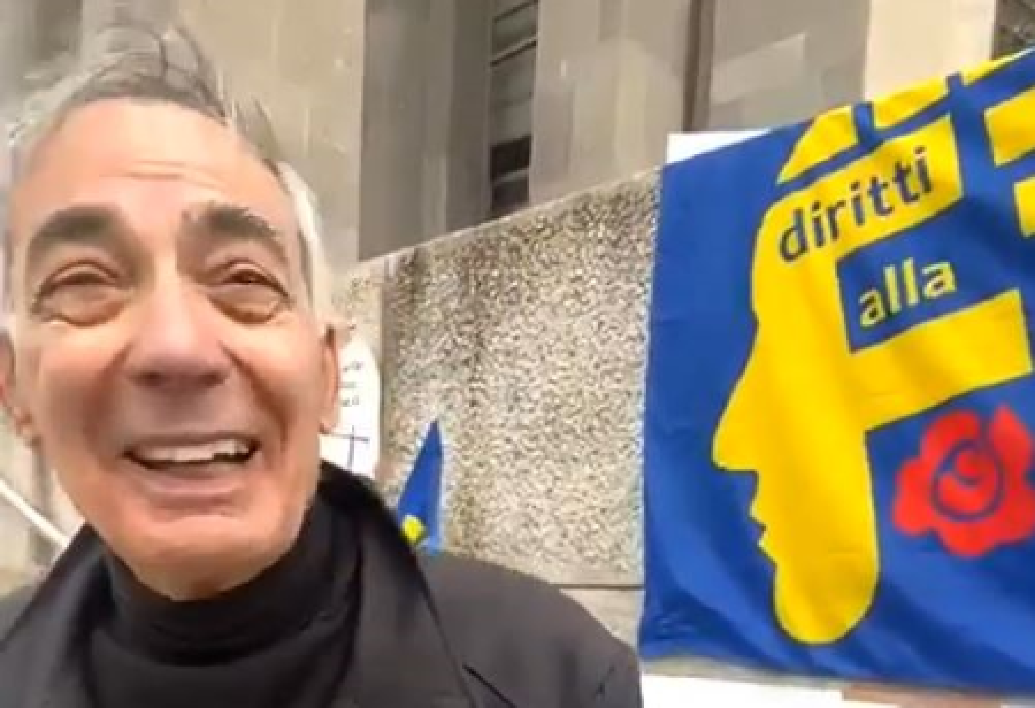 Sorride il professor Gigi Monello, dopo essere stato assolto con formula piena dalla falsa accusa di maltrattamento nei confronti della madre. Dietro di lui la bandiera di Diritti alla Follia, l’Associazione che lo ha sostenuto in tutta la vicenda processuale